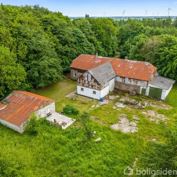 En gammel gård med rustne tage står omgivet af tæt skov. Bygningen har flere sammenhængende sektioner, og der er en lille strukturel tilbygning. Græsplæne og træer dominerer omgivelserne.