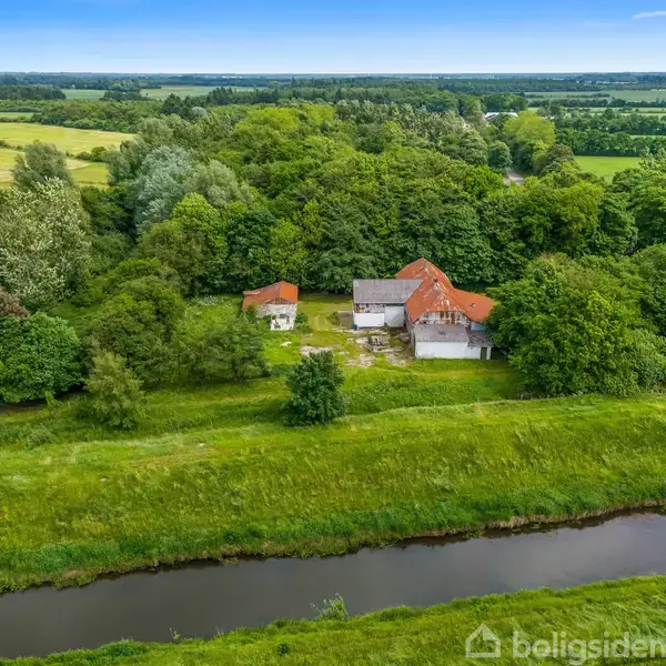 En landskabsscene viser en gård med tre røde tegltagsbygninger omgivet af tæt skov. Bygningerne ligger nær en flod, omgivet af grønne marker og skove under en åben himmel.