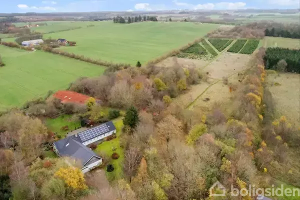 Hus med solpaneler ligger i frodigt grønt område omgivet af marker og træer, bygninger spredt i landskabet, under en delvist overskyet himmel.