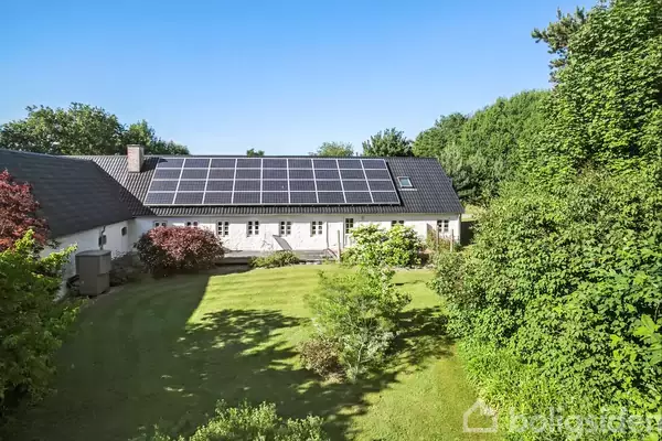 Et hvidt hus med solpaneler på taget står i en velplejet have omgivet af frodige grønne træer. Solen skinner klart på græsplænen og buskene.