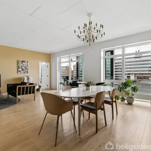 Spisebord med stole står i en moderne stue. Omkring er der en sofa, et TV på et stativ, og store vinduer med udsigt til bygninger.