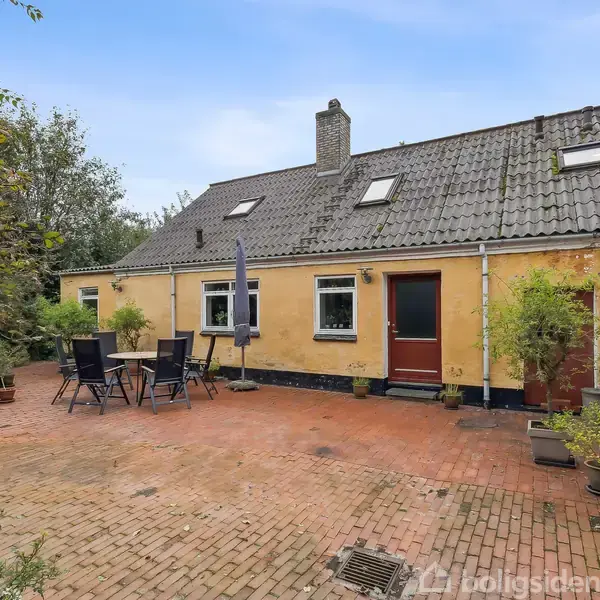 En gul murstensbygning har en gårdhave med røde fliser, borde og stole under en parasol. Træer og buske omgiver området, som udstråler en hyggelig landlig atmosfære.