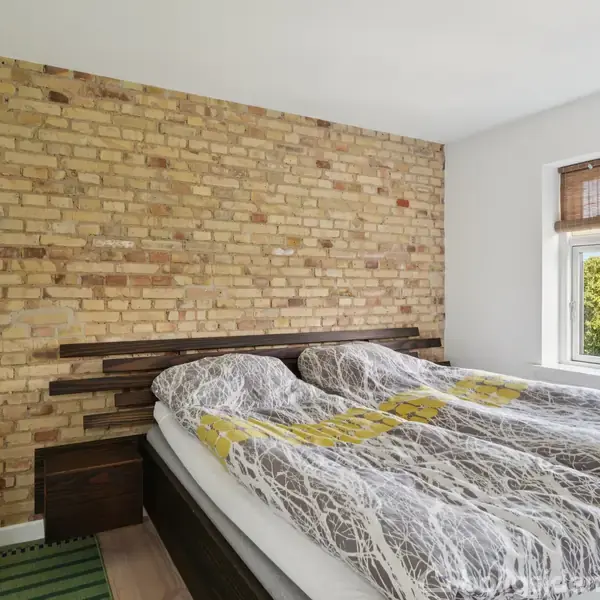 Seng med mønstret sengetæppe står mod en murstensvæg i et lyst soveværelse. Ved siden af er et vindue med udsigt til grønne træer.