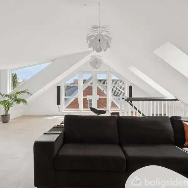 Sort sofa står i stue, med skrå vægge og store ovenlysvinduer. En hvid lampe hænger i loftet, og en plante står ved det åbne vindue.