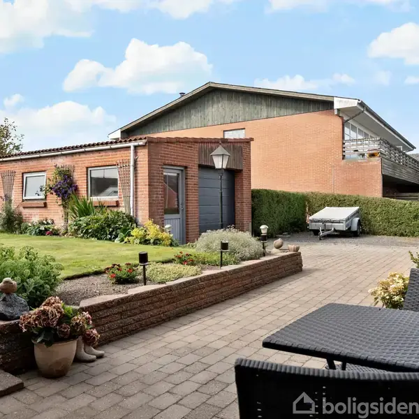 Terrasse med fliset gulv og sorte møbler. Til venstre trappe, blomster og en græsplæne. Rødt murstenshus med carport i baggrunden. Solrig dag med spredte skyer.