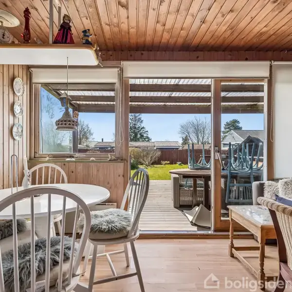 Rundt spisebord med stole står ved store vinduer, der åbner ud til en terrasse og have. Omkring bordet er der trævægge og dekorative tallerkener.