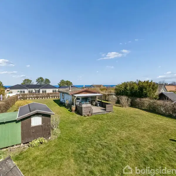 Hus med have og skur, set udefra. Haven er stor med græsplæne. Skuret er grønt og brunt. I baggrunden ses flere huse og træer samt en klar blå himmel.