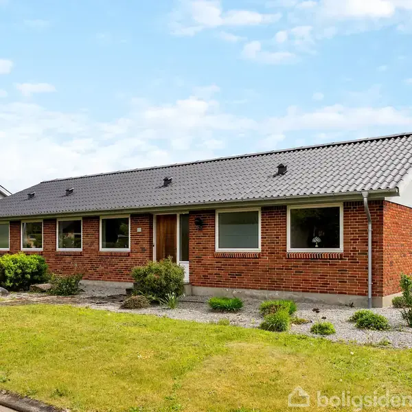 Et murstenshus med sort tag står midt i en velholdt have med græsplæne og buske. Et "Til Salg" skilt ses i forhaven. Himlen er blå med få skyer.