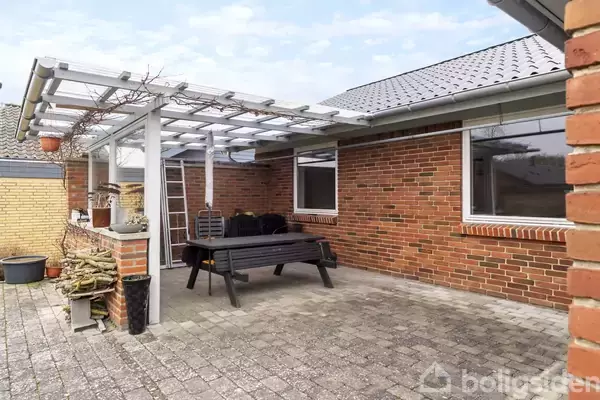 En terrasse med et bord og bænke omkranset af murede vægge, overdækket med en pergola i en gårdhave. Fliser dækker jorden, brændestabel og planter i krukker langs muren.