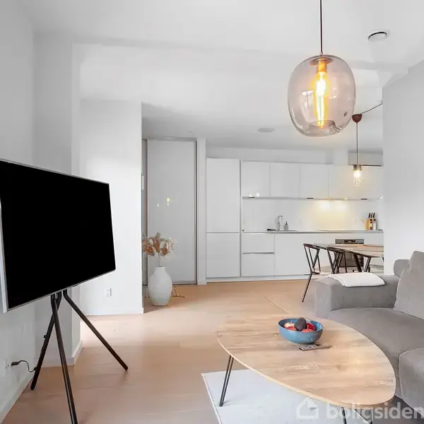 TV står på stativ, viser intet indhold; i den moderne stue er der en sofa, træbord med skål. I baggrunden ses køkken med hvide skabe og spisebord med stole.