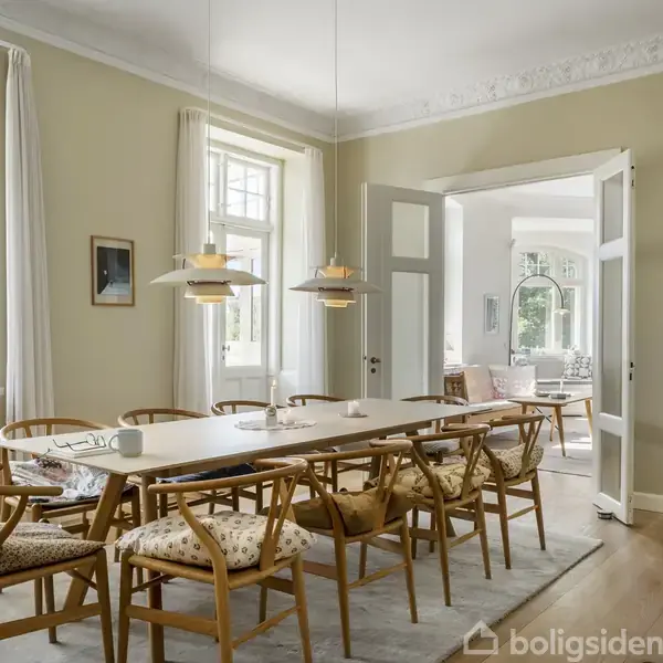 Et moderne køkken med grå skabe og stålovn. Over ovnen hænger en stål-emhætte. Bordpladen er dækket med køkkenredskaber, planter og dekoration. Sollys strømmer ind gennem et stort vindue.