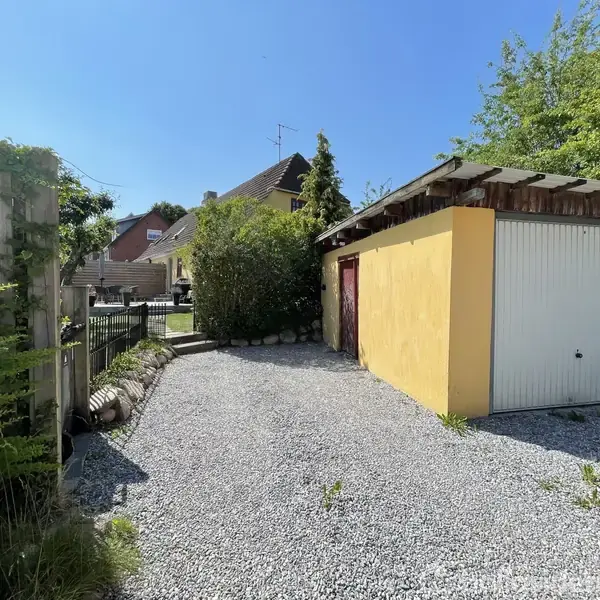 En gul garagebygning med en hvid port står i en grusbelagt indkørsel. Omkring bygningen er der grøn vegetation, og bagved ses et villakvarter med huse og stakitter.