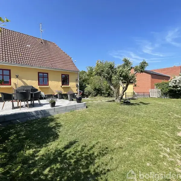 Et gult hus med rødt tegltag står ved en terrasse med havemøbler. Haven er græsbevokset og har træer, i et solrigt område med blå himmel.