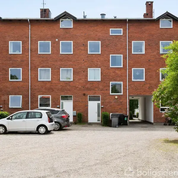 En tre-etagers murstensbygning med firkantede vinduer, står over for en parkeringsplads med to biler. En grøn busk er til højre i billedet.