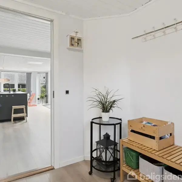 Indgang med en bænk fyldt med en trækasse; en plante står på et smalt, rundt bord; i baggrunden ses et lyst, åbent køkkenområde med møbler.