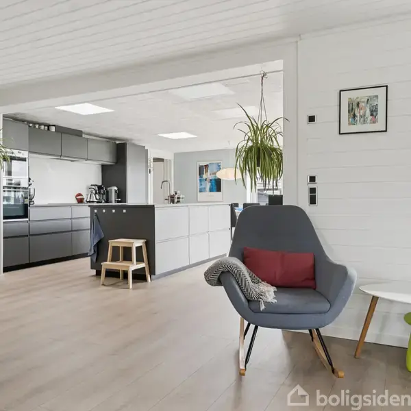 En grå lænestol med pude og tæppe står i en lys stue ved siden af et lille bord; moderne køkken ses i baggrunden med hængende planter og en bænkeskammel.