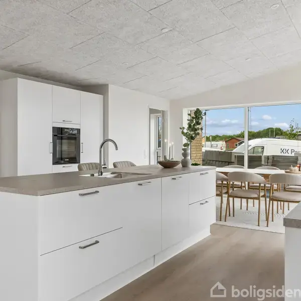 Et moderne køkken med hvid køkkenø, indbygget ovn og spiseplads. Store vinduer bagest giver udsigt til grønt område og huse udenfor. Bordet er dækket med dekorative genstande.