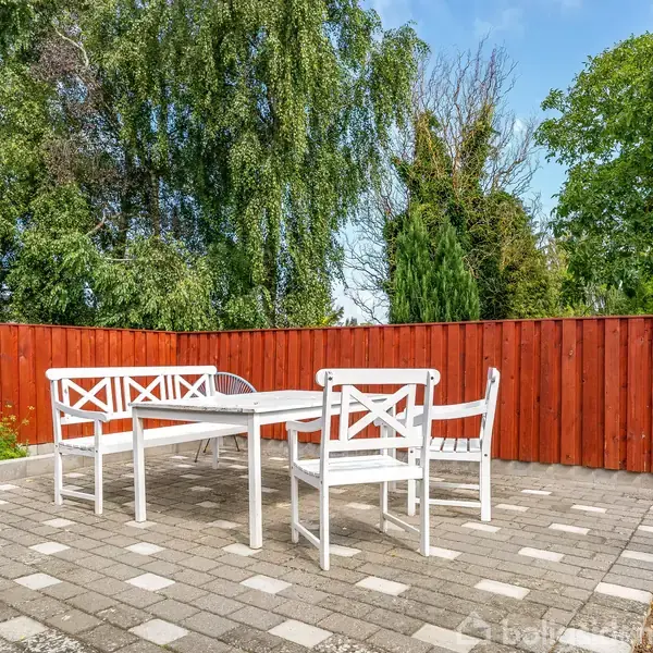 Hvide havemøbler står på en flisebelagt terrasse omgivet af en rød træhegn. Baggrunden viser frodige grønne træer, der skaber en indbydende udendørs atmosfære.