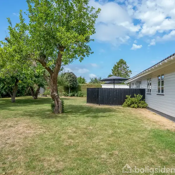 Et hvidt hus står på en velplejet græsplæne med træer i nærheden. En sort terrasseparasol ses bag et hegn. Skyer dækker delvist den blå himmel.