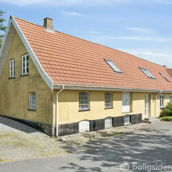 Gul landhusbygning med rødt tegltag og flere ovenlysvinduer. Bygningen står stille på en grusvej omgivet af træer og grønne buske under en blå himmel.