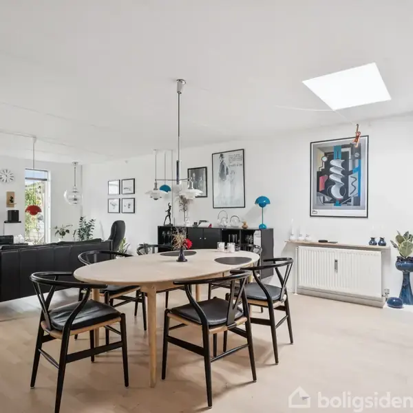 Et moderne spisebord med sorte stole står i en lys stue. Omkring bordet er der diverse møbler, kunst på væggene, og en fjernsynsskærm viser et natursceneri.