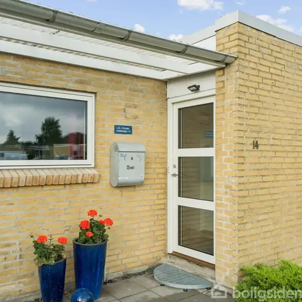 Et gult murstenshus med en glasdør og postkasse ved siden af. Blomster i blå potter står foran vinduet. Indkørslen er flisebelagt. Teksten lyder: "GLS Pakkeshop 5520", "14".