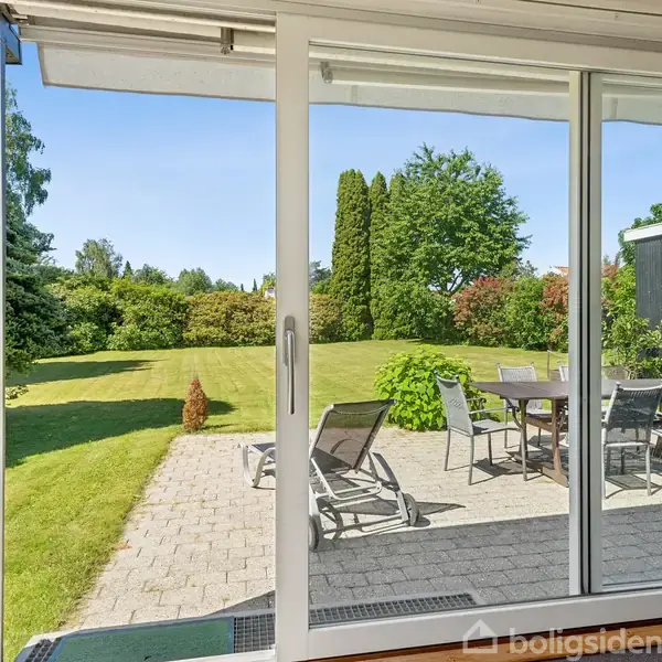 Skydedør åbner til terrasse med havemøbler, beliggende i en velplejet have med grøn græsplæne og omgivende buske og træer under klar blå himmel.