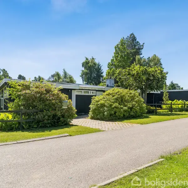 Et moderne hus omgivet af frodige grønne buske og træer står ved en stille vej. Indkørslen er brolagt, og der er en garage på siden.