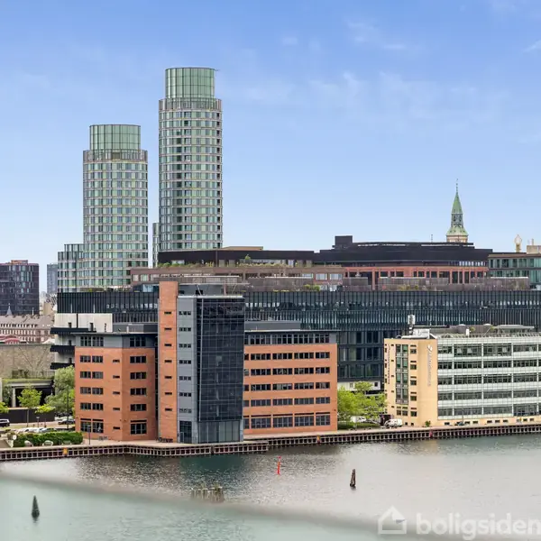 Flere moderne bygninger spejler sig i en rolig vandkanal. I baggrunden hæver højhuse og en kirkespir sig mod den klare blå himmel.