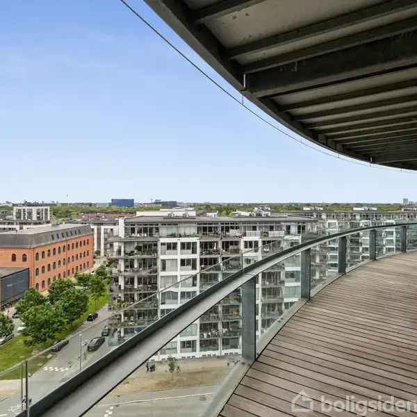 En buet altan lavet af træ, der strækker sig langs en moderne bygning i en bygrænse, med udsigt over andre bygninger og grønne områder under en klar himmel.