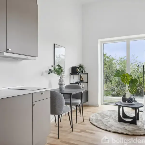 Et moderne køkken-alrum med grå skabe, en rund spisebord med to stole, store vinduer med udsigt til en grøn have, og en sofa med puder i et lyst rum.