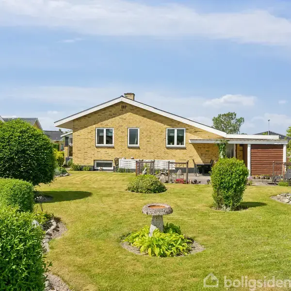 Et gult murstenshus står stille i en velplejet have med grønne buske og en græsplæne. Der er en stenterrasse foran huset under en blå himmel.