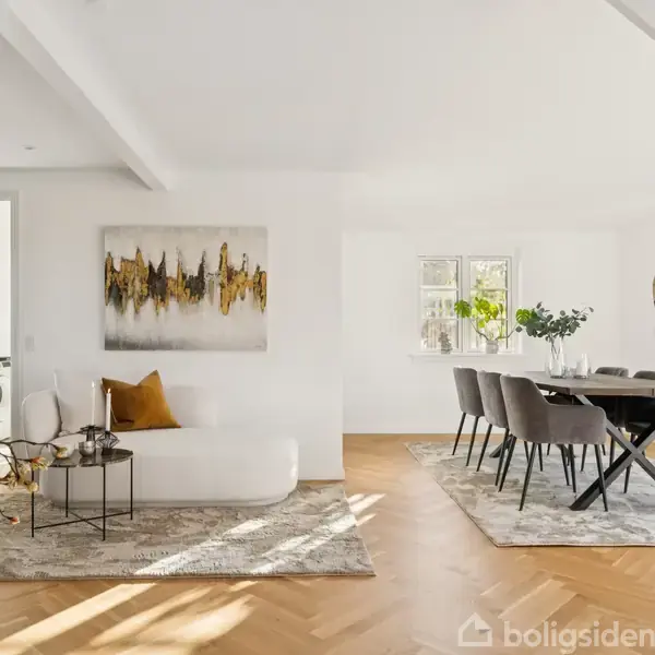 Sofa med pude står i sollys ved siden af kunst på væggen; spisestue med bord og stole til højre; store vinduer slipper lys ind. Ingen tekst.