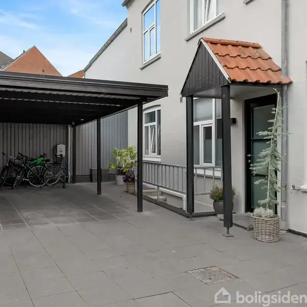 Carporten rummer flere cykler ved siden af en hvid bygning med et lille udhæng over hoveddøren. Fliser dækker gårdspladsen, og nogle planter ses langs væggen.