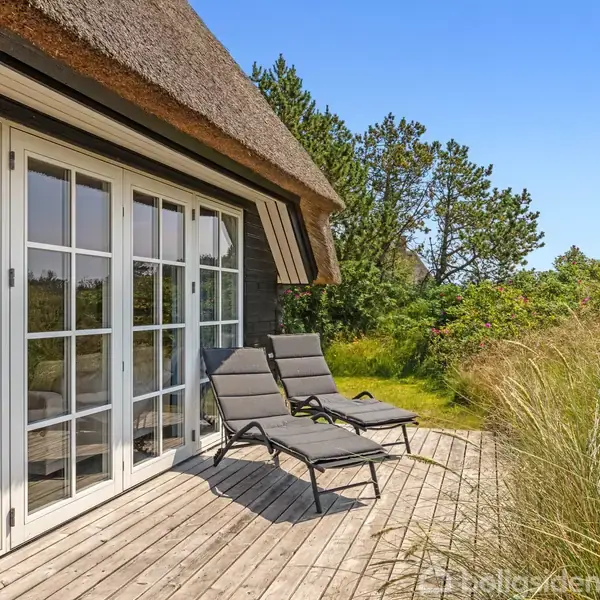 To liggestole står på en træterrasse ved et hus med stråtag. Husets store glasdøre åbner ud mod en grøn have fyldt med høje græsser og træer under en klar blå himmel.