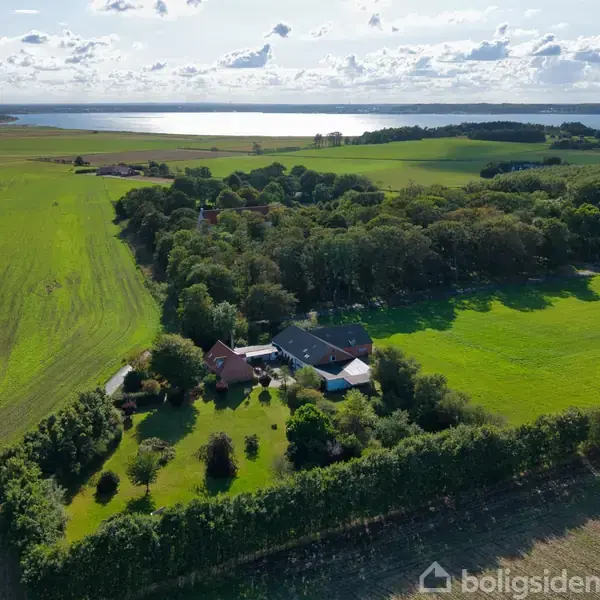 Hus omgivet af haver og træer ligger i frodigt landbrugsområde, med udsigt til sø i det fjerne under en skyet himmel.