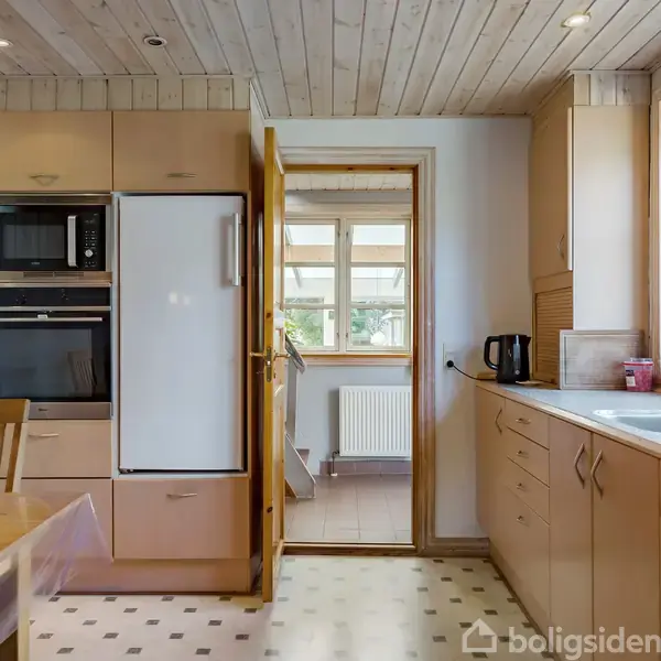 Et køkken med træskabe og hvidevarer som indbygget ovn og køleskab. En skål med frugt står på bordet. Gennem en dør ses et tilstødende rum med radiator og vinduer.