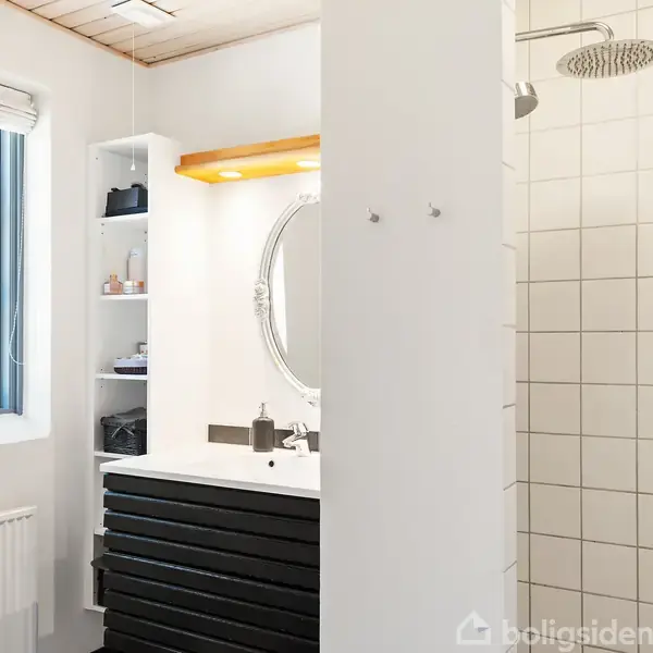 Vask med sort skab nedenunder, spejl og lys over. Til højre en bruseniche med fliser. Til venstre et vindue og radiator. Skiftevis lyse og mørke elementer skaber kontrast.