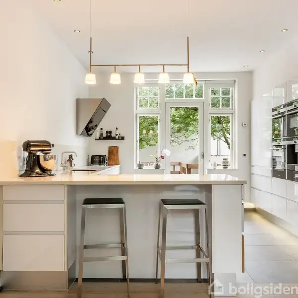 Et moderne køkken med en hvid ø-bord, to metalstole og hvide skabe. En kaffemaskine står til venstre. Baggrunden viser store vinduer med udsigt til grønne træer.