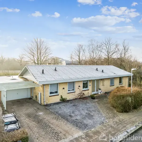 En enkeltstående etplanshus med gult murværk står stille i et vinterlandskab. Omgivet af grusbelagt indkørsel og en garage. Træer uden blade ses i baggrunden.