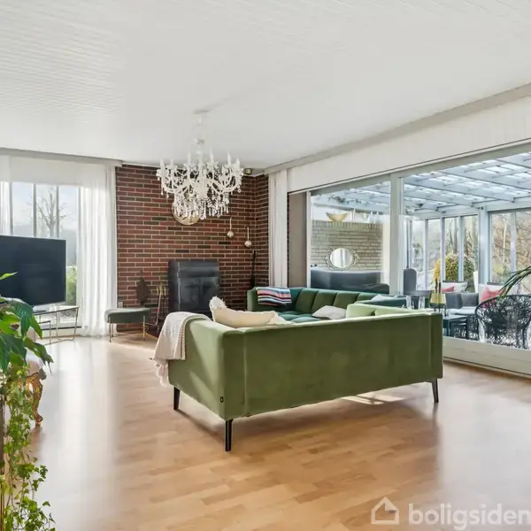 Grøn sofa står på trægulv foran pejs i lys stue med store vinduer. En lysekrone hænger over sofaen, og flere grønne planter dekorerer rummet.