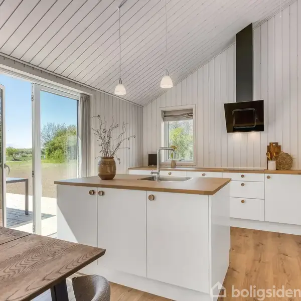 Et moderne køkken med hvide skabe og en træbordplade. Det står i et lyst rum med store vinduer, der åbner ud til en terrasse med udsigt over grøn natur.