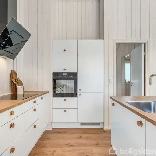 Køkken med hvide skabe og træbordplader; en indbygningsovn i midten, en sort, skrå emhætte til venstre, og vask med høj hane til højre. Mørkt trægulv.
