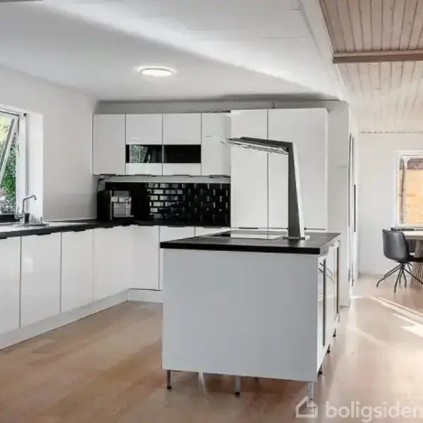 Et moderne køkken med hvide skabe og en sort bordplade. En køkkenø har en indbygget vask. I baggrunden ses et spiseområde med et rundt bord og stole ved et vindue.