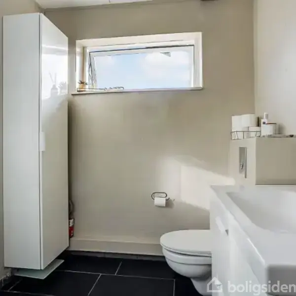 Badeværelse med hvid håndvask og sort vandhane, væghængt toilet, og høj skab. Et lille vindue slipper lys ind. Gulvet er sort, og der er en brusekabine til venstre.
