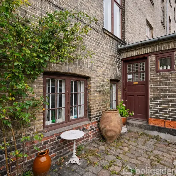 En murstensbygning med en brun dør, store vinduer og klatrende planter. Flasker står i vindueskarmen. En lille gårdhave med en rund hvid metalbord og to store terracotta-krukker.