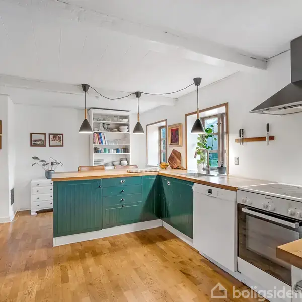 Et køkken med grønne skabe, en ovn og opvaskemaskine, placeret i et lyst rum med trægulv. Over bordpladen hænger tre sorte lamper, og vinduerne giver naturligt lys.