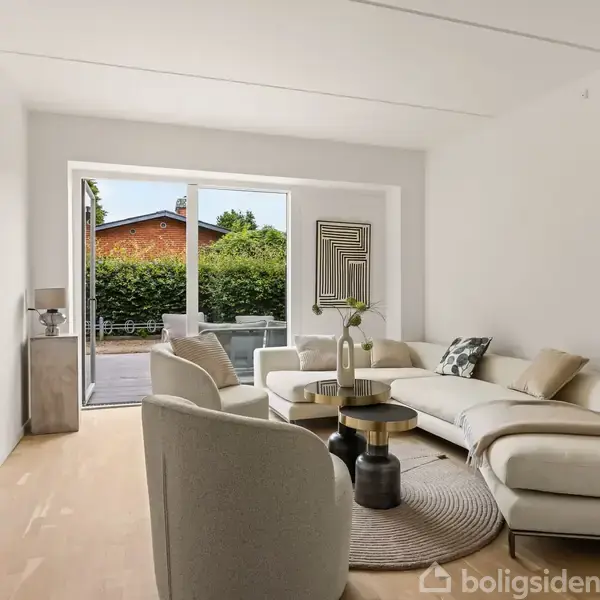 En lys stue med moderne møbler; en cremefarvet sofa og stole står omkring små, sorte borde. Rummet har trægulv, et tæppe, og glasdøre åbner til en terrasse med hæk.