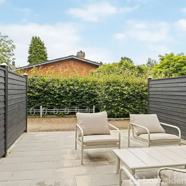 Terrassemøbler står på en flisebelagt gårdhave, bestående af to stole og et bord. Omkring er der mørke træhegn og frodige buske; i baggrunden ses et rødt murstenshus.