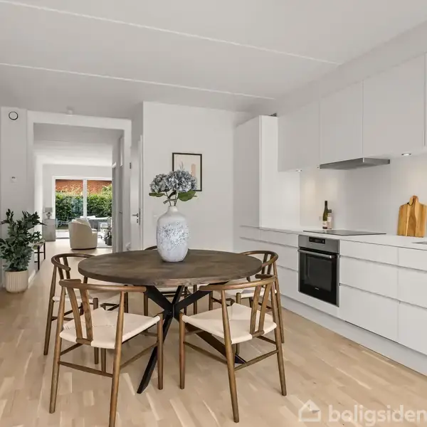 Rundt spisebord med fire stole står i et lyst køkken-alrum. Køkkenet har hvide skabe, indbygget ovn og vask. Foran er en vase med blomster. I baggrunden ses en åben stue.